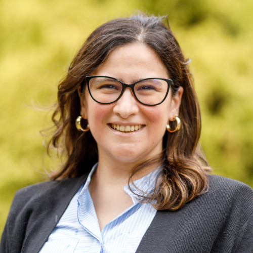Faculty Sara Machado smiling at the camera wearing glasses and a gray cardigan with green foliage in the background.