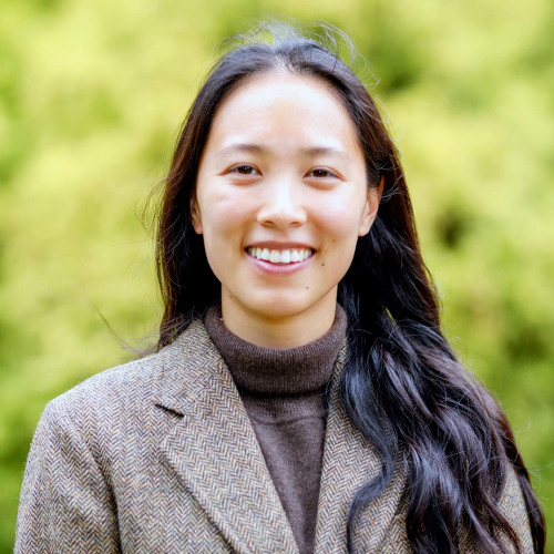 Headshot of faculty Olivia Zhao, wearing a brown herringbone blazer with green foliage in the background.