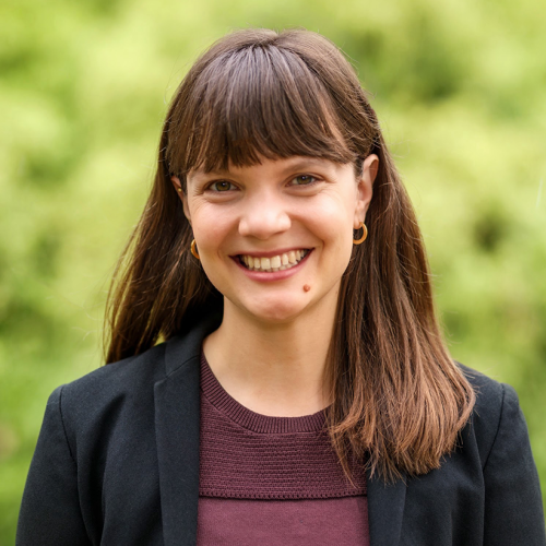 Headshot of faculty member Haley Sullivan smiling at the camera, wearing a black blazer, with green foliage in the background