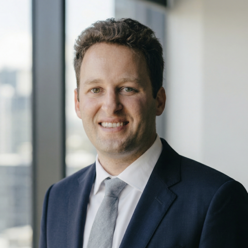 Headshot of faculty member Daniel Arnold, a man with short, wavy brown hair, smiling in a dark blue suit, white collared shirt, and grey tie. He is standing indoors in front of a window with a blurred city view.