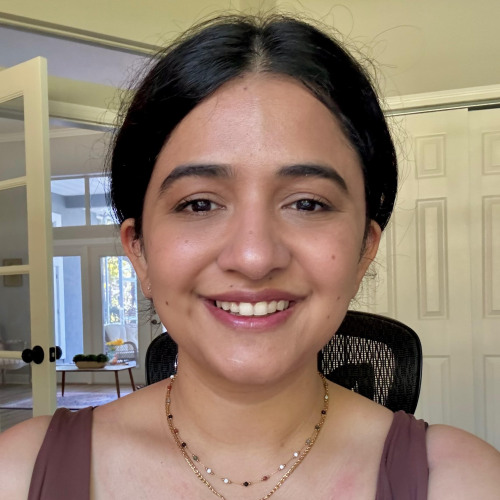 Headshot of faculty member Mah-Afroze Chughtai smiling warmly at the camera, wearing a mauve top and a double-layered gold necklace with a green pendant, seated in a bright indoor room.