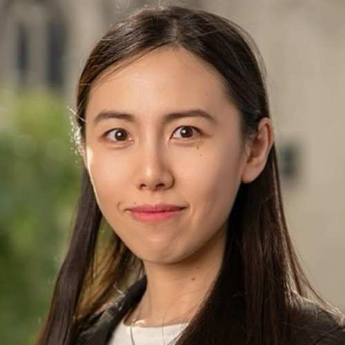 Headshot of faculty member Xiaoxi Zhao smiling, wearing a black blazer, white top, and a silver necklace, against a softly blurred outdoor background.