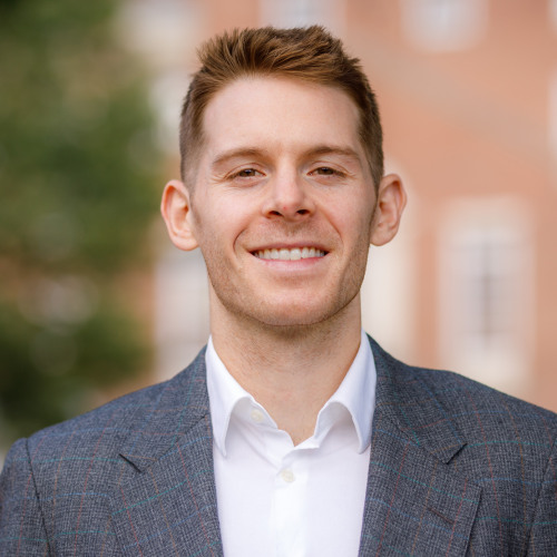 Headshot of faculty Hayden Rooke-Ley smiling at the camera in a gray suit in front of foliage and city buildings.