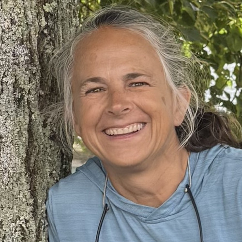 Headshot of faculty member Gaelen Adam smiling while wearing a blue shirt with a tree in the background.
