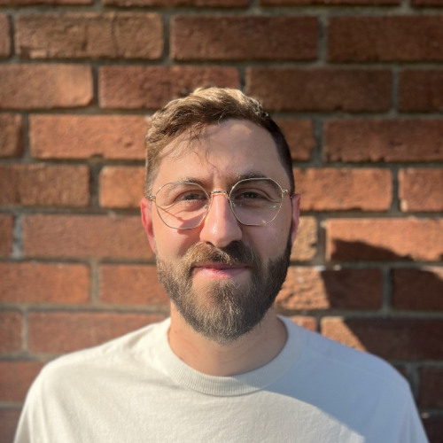 Headshot of doctoral student Josh Weiner, smiling at the camera in a white crewneck shirt, wearing glasses, against a brick background.