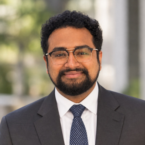 Doctoral student Avirath Vaidya smiling at the camera wearing a dark gray suit and glasses, with foliage and a building in the background.