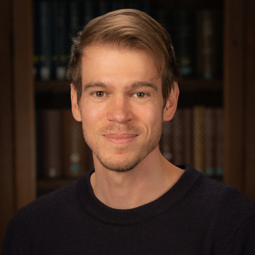Headshot of faculty member Olivier Wouters smiling, wearing a dark collared shirt against a bookshelf background.