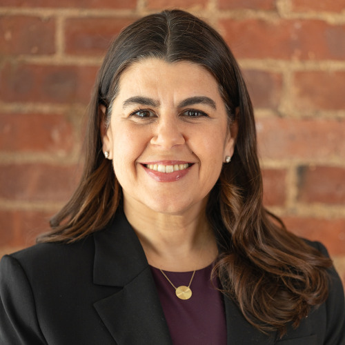 Headshot of faculty member Dara Kass, wearing a purple shirt and black blazer, smiling against a brick background.