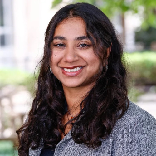 Headshot of doctoral student Vani Agarwal smiling at the camera in a textured blue blazer with foliage in the background.