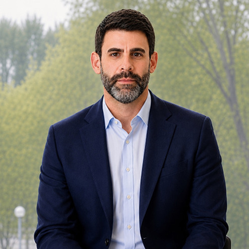 Headshot of faculty member Matthew Vogel wearing a navy blue blazer, slightly smiling at the camera with a view of trees through a window in the background.