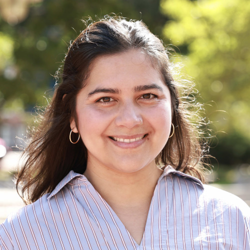 Headshot of doctoral student Dea Oviedo wearing a striped dress shirt, smiling at the camera against foliage and cityscape.