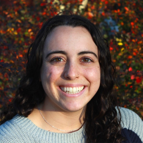 Headshot of doctoral student Bekka Stein wearing a light blue sweater, smiling at the camera with foliage in the background.